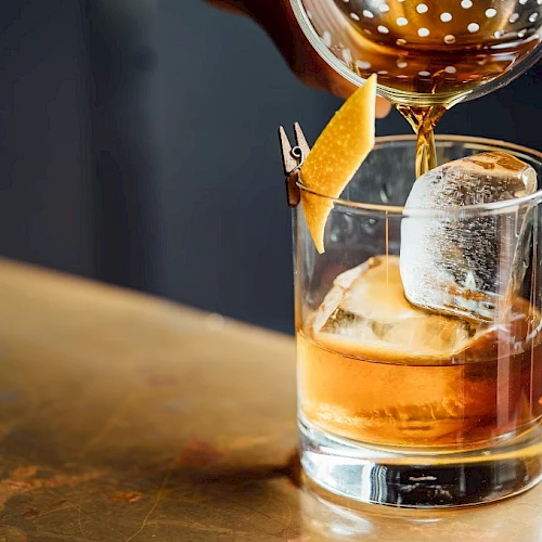 A glass of whiskey on the rocks with a slice of orange garnish, being topped up from a shaker above.