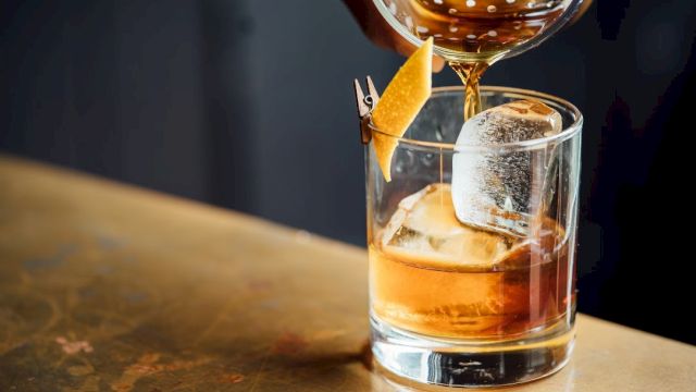 A glass of whiskey on the rocks with a slice of orange garnish, being topped up from a shaker above.