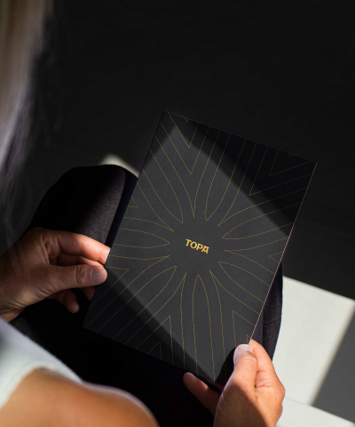 A person holds a black notebook with a gold sunburst design and the word &ldquo;TOPA&rdquo; in the center, outdoors on a sunny day.