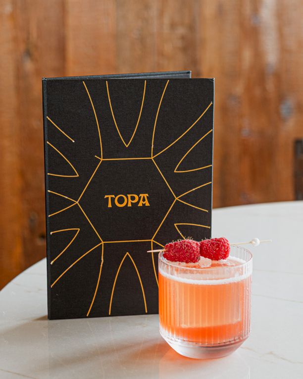 A black TOPA gift box sits beside a pink cocktail in a glass topped with raspberries.