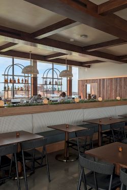 A stylish bar and dining area with wooden beams, a long counter with stools, and tables set for guests, backdrop of bottles and windows, cozy lighting.
