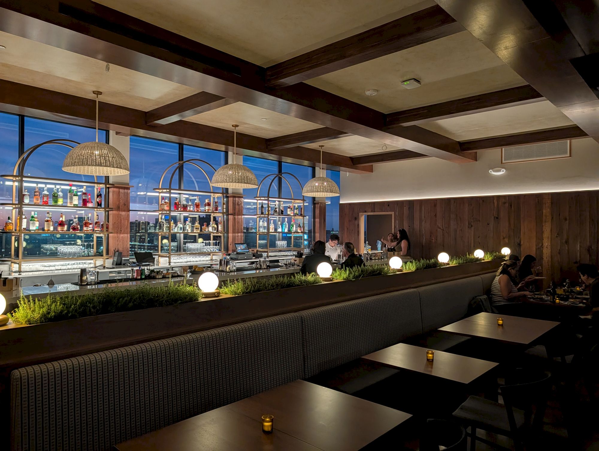 A stylish hotel lounge or restaurant with dark wood accents, a long cushioned bench, small tables, warm lighting, and a bar area by large windows.