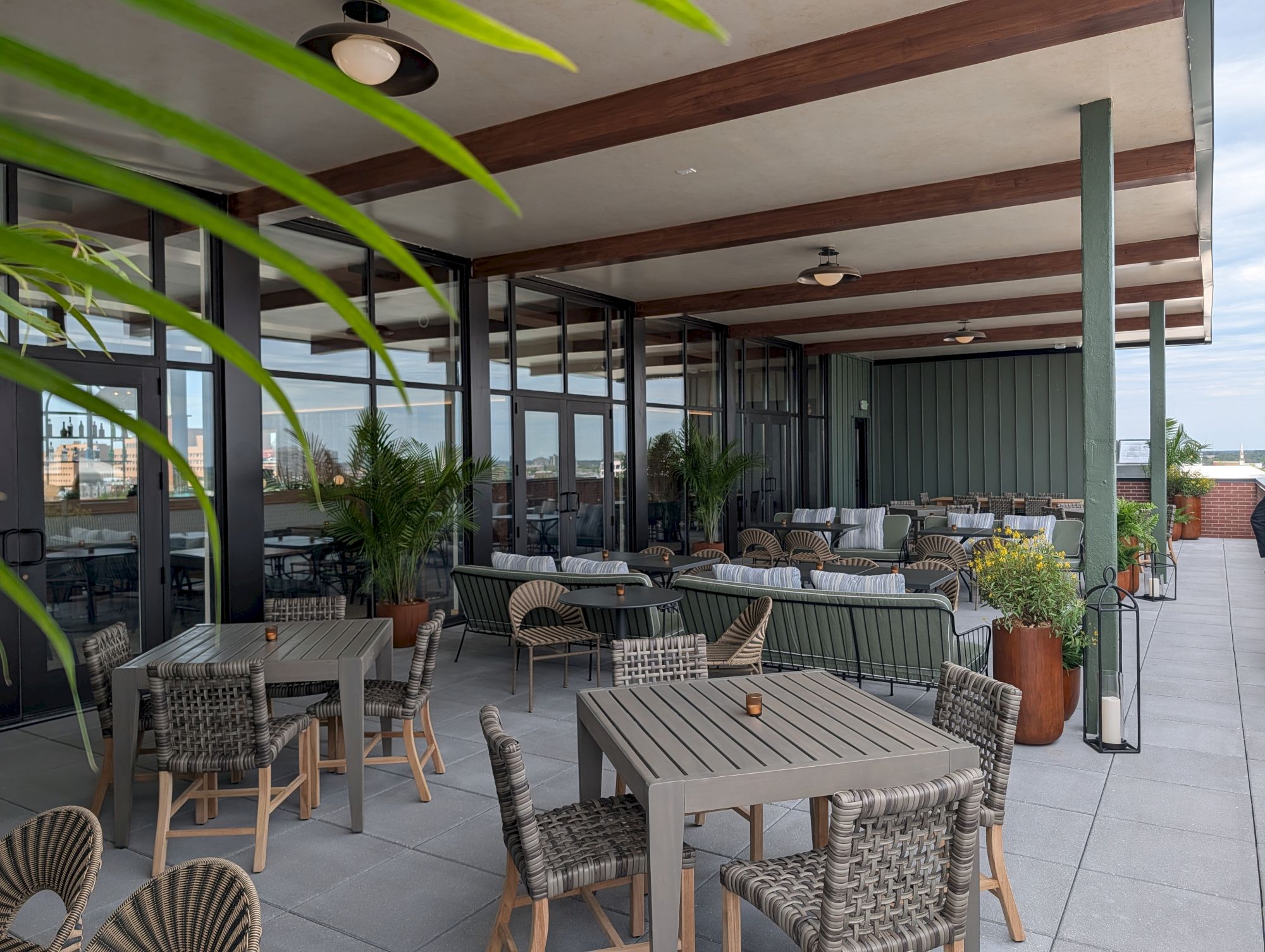 A spacious outdoor dining area with woven chairs, wooden tables, potted plants, and a covered patio under a wooden beam roof.