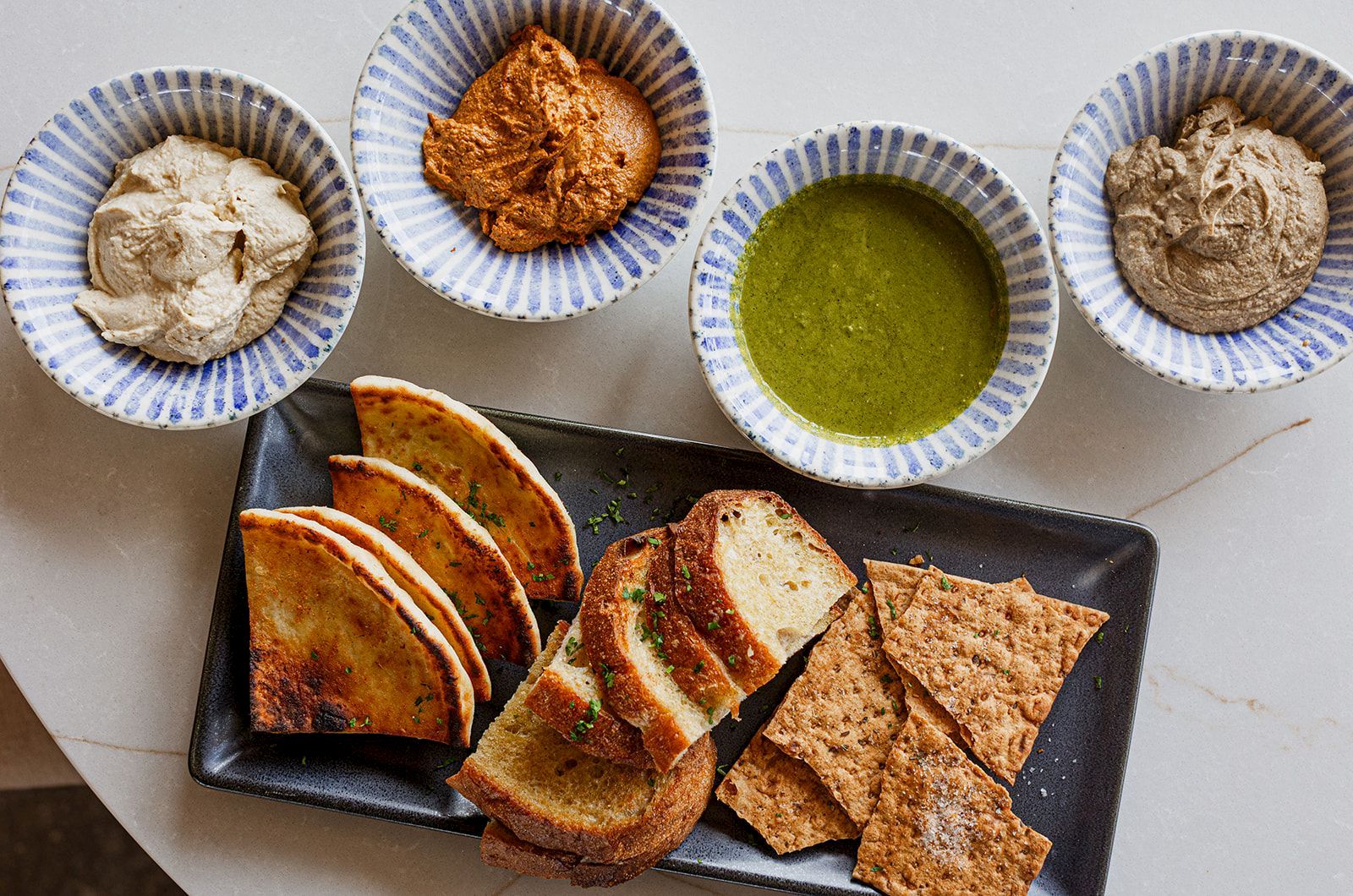Platter of grilled bread slices and crackers with hummus, a meat dip, and green herb oil in small bowls.