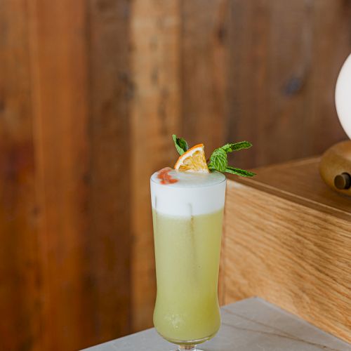 A tall pale-yellow cocktail in a glass, topped with mint and a lemon slice, served on a wooden table.