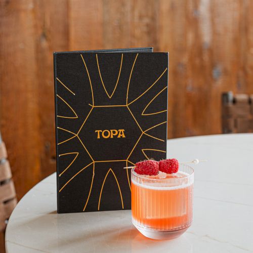 A black TOPA box and a pink cocktail topped with raspberries sit on a marble table.