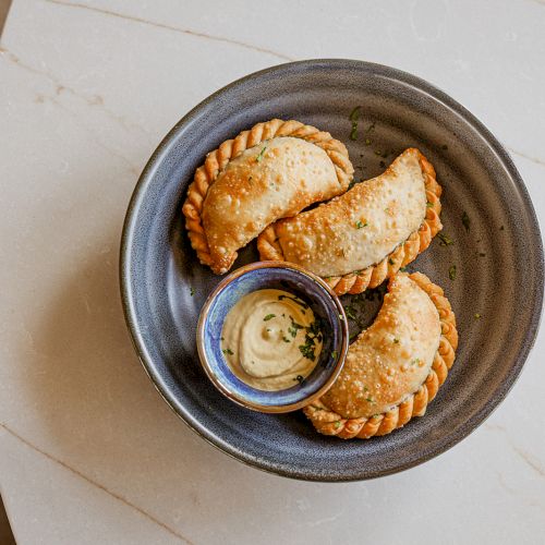 A plate of baked empanadas served with a small cup of creamy dipping sauce and green herbs for garnish.