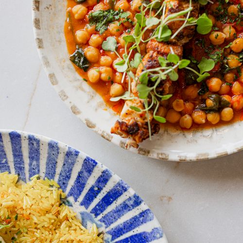 A bowl of spicy tomato chickpea stew with herbs and sprouts, next to a striped plate of yellow basmati rice.