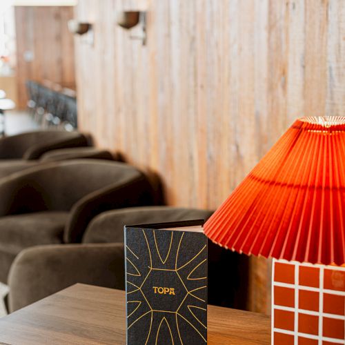 A rustic lounge scene with a wooden side table, a black gift box, an orange lampshade, and a textured wooden wall backdrop.